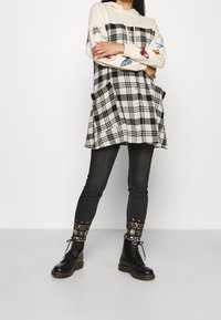 Persona que lleva una sudadera color crema con diseños de mariposas y abejas, un chaleco de cuadros en blanco y negro, jeans negros con bordados florales y botas negras con cordones.