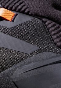 Chaussures noires en tissu texturé avec des rayures verticales, des accents en caoutchouc et une sangle orange contrastante.