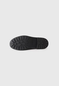 Suela de zapato de goma negra con un patrón de banda de rodadura profundo que incluye tacos rectangulares y en forma de estrella, mostrada sobre un fondo blanco liso.