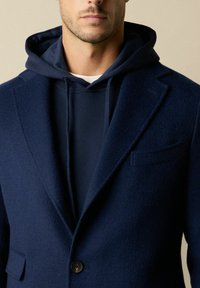Granatowa marynarka z mieszanki wełny na granatowej bluzie z kapturem o dopasowanym kolorze. Marynarka ma klapę z wycięciem, zapięcie na jeden guzik oraz kieszeń na wysokości klatki piersiowej.