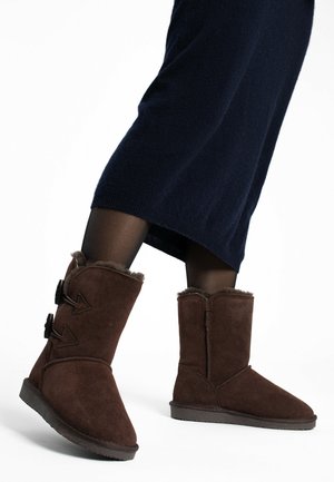 Brown suede ankle boots with a soft fur lining, angled design, dual side buttons, and a flat rubber sole. Worn with a dark-knit skirt.