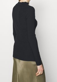 Haut noir à manches longues côtelé avec encolure à volants. Coupe ajustée, tissu texturé. Porté avec une jupe en satin vert olive clair, partiellement visible.