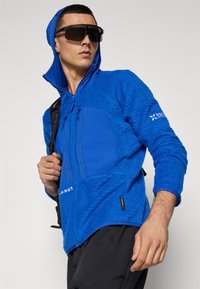 Jachetă din fleece albastră, cu țesătură texturată, glugă și buzunar frontal cu fermoar. Prezintă un logo pe mânecă și este asortată cu pantaloni negri.
