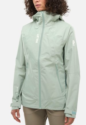 Femme portant une veste imperméable vert clair avec fermeture éclair frontale, capuche et poches zippées, debout devant un fond uni.