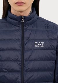 Navy puffer jacket with quilted design, high collar, and full front zipper. Features embroidered EA7 logo on the chest. Smooth, shiny texture.