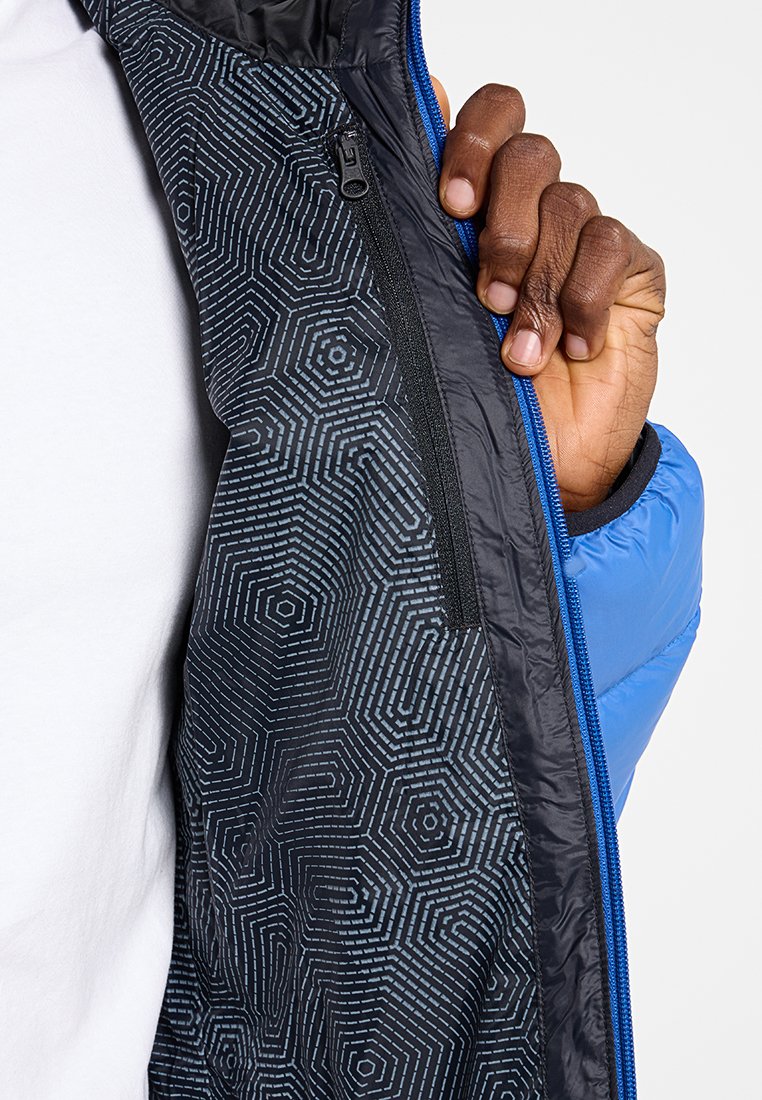 Main tenant une veste bleue ouverte montrant une doublure intérieure noire avec un motif géométrique hexagonal et une poche zippée verticale.