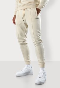 Cremfarbene Jogginghose mit elastischen Bündchen, Bindegurt an der Taille und Seitentaschen. Schwarzes Branding-Detail am Oberschenkel. Kombiniert mit weißen Turnschuhen.