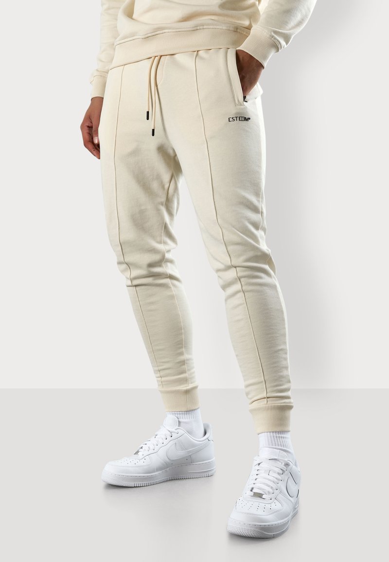 Cremfarbene Jogginghose mit elastischen Bündchen, Bindegurt an der Taille und Seitentaschen. Schwarzes Branding-Detail am Oberschenkel. Kombiniert mit weißen Turnschuhen.