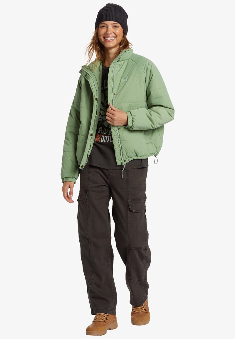 Giacca a vento verde con chiusura a zip e bottone, polsini elasticizzati, abbinata a pantaloni cargo neri e stivali beige, completata da un berretto nero.