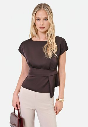 Femme blonde portant une blouse marron foncé nouée à la taille et un pantalon beige clair, tenant un sac à main marron foncé et portant un bracelet en or.