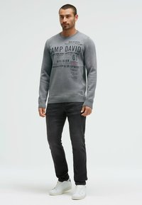 Grauer Pullover mit aufgedrucktem Text in verschiedenen Schriftarten, gerippten Bündchen und Saum. Kombiniert mit dunklen Jeans und weißen Sneakers. Einfaches, lässiges Outfit.