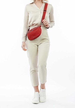 Borsa a tracolla rossa in pelle con forma curva e accessori in argento, indossata sopra una camicia beige e pantaloni crema, abbinata a sneaker bianche.