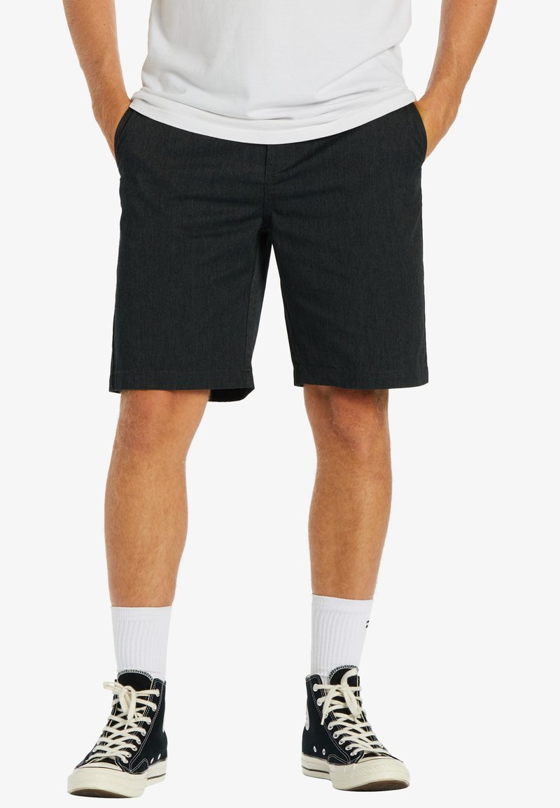 Uomo che indossa pantaloni corti neri, calze bianche fino al polpaccio, sneakers alte nere e una t-shirt bianca con le mani nelle tasche.