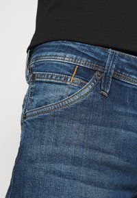 Närbild av en jeansficka i blå denim med orange sömmar och en svart topp instoppad.