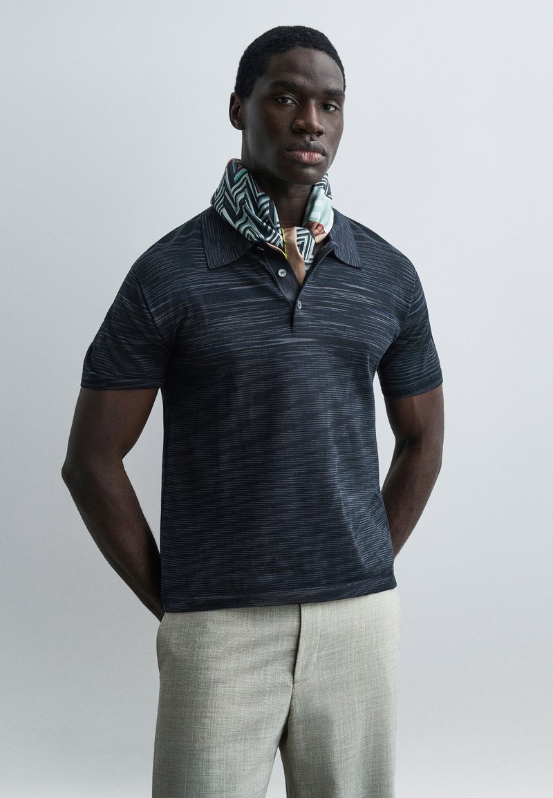 Άντρας που φοράει σκούρο κοντομάνικο πόλο με σχέδια, ανοιχτό μπεζ παντελόνι και πολύχρωμο μαντήλι με σχέδια γύρω από το λαιμό του.