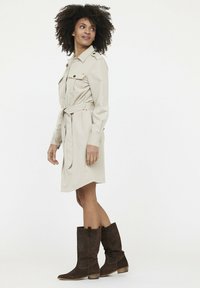 Beiges Hemdkleid mit langen Ärmeln, Knopftaschen und einem gebundenen Taillenbund. Kombiniert mit braunen Wildlederstiefeln mit mittlerer Schafthöhe und strukturiertem Design.