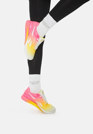 Nogi w czarnych legginsach, noszące białe skarpetki Reebok oraz różowo-żółte buty do biegania Reebok na białym tle.