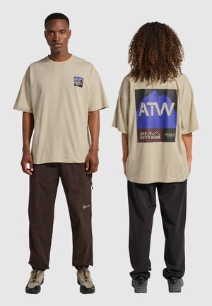 Beige übergroßes T-Shirt mit einem blauen und schwarzen Grafikdesign auf dem Rücken, aus weichem Baumwollstoff, kombiniert mit dunklen Cargo-Hosen.