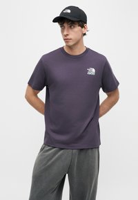 Νεαρός άνδρας με μαύρο καπέλο και σκούρο μωβ μπλουζάκι The North Face με λογότυπο, στέκεται με τα χέρια πίσω από την πλάτη σε απλό φόντο.