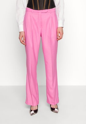 Frau trägt hochtaillierte, locker sitzende rosa Hose mit Falten, weiße Hemdmanschetten und schwarze spitze High Heels mit metallischen Spitzen.