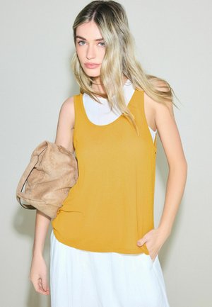 Junge Frau mit langem blondem Haar, die ein ärmelloses, senfgelbes Oberteil über Weiß trägt und eine hellbraune Wildledertasche vor beigem Hintergrund hält.