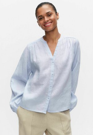 Smilende kvinde med håret trukket tilbage, iført en lyseblå knapbluse og beige bukser, stående med hænderne i lommerne mod en hvid baggrund.