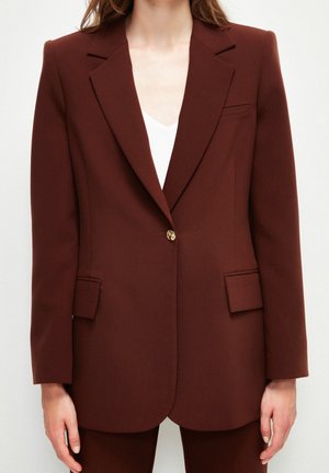 Vrouw draagt een op maat gemaakte donkerbruine blazer met een enkele gouden knoop, revers met inkeping, klepzakken en een wit shirt eronder.