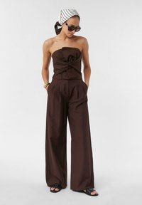 Braunes trägerloses Top mit Knoten-Detail, kombiniert mit hochtaillierten, weiten Hosen. Das Outfit hat eine glatte Textur. Schwarze Sandalen runden den Look ab.