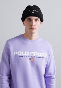 Svetlo fialová mikina s nápisom "POLO SPORT RALPH LAUREN" a americkou vlajkou. Čierna čiapka s nápisom "POLO SPORT". Hladká textúra.