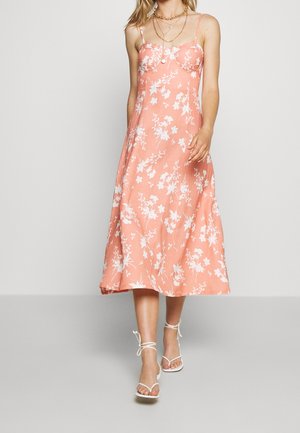 Kvinde iført en koralrosa midi-kjole med hvidt blomsterprint, tynde stropper og hvide sandaler med stropper, gående mod en ensfarvet baggrund.