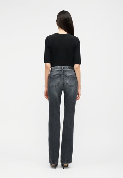 Vrouw met lang donker haar, gekleed in een zwart topje met korte mouwen, hooggetailleerde verweerde zwarte spijkerbroek en zwarte hoge hakken, gezien vanaf de achterkant.