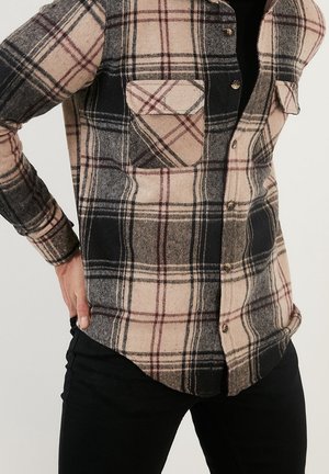 Man draagt een beige, zwart en rood geruit overhemd met knopen en twee borstzakken, gecombineerd met een zwarte broek, hand rustend op de heup.