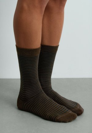 Jambes portant des chaussettes marron mi-mollet avec de fines rayures horizontales noires, debout sur un sol gris lisse sur un fond clair uni.