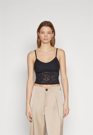 Musta pitsi crop top ohuilla olkaimilla, jossa on kukkakuosi ja hampaat reunoissa, yhdistettynä korkea vyötäröisiin beigeihin housuihin.