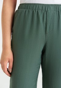 Gröna, lätta, elastiska byxor med midjeresår som har en slät textur och subtila sömnadsdetaljer, med en närbild av tyget och passformen.