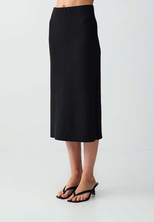 Woman wearing a black midi skirt paired with black heeled sandals with thong straps, standing against a plain light background.