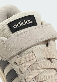 Beige sneaker met suède accenten, een mesh-tong en een zwart Adidas-logo. Beschikt over lichtgrijze strepen en geweven veters.