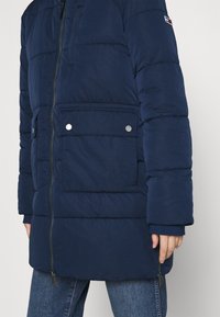 Veste matelassée bleu marine avec fermeture éclair, deux poches avant à bouton-pression, poignets côtelés et texture matelassée. Détail du logo sur la manche.