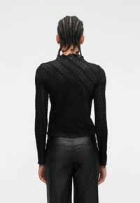 Svart langermet topp med et lacy, teksturert mønster og høy hals. Båret med svarte skinnbukser, som viser en tettsittende silhuett.