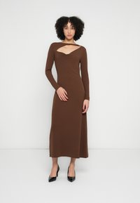 Brūnas krāsas kleita ar garām piedurknēm, V veida izgriezumu, gludu tekstūru, piegulošu siluetu un potītes garuma apakšu, kombinēta ar melniem augstiem papēžiem.