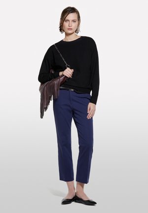 Donna che indossa un maglione nero, pantaloni cropped blu navy, ballerine nere e porta una borsa a tracolla marrone con frange e catena.