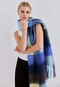Wollen sjaal met een mix van blauw, grijs en crèmekleurige ruitpatronen, met franjes aan de randen, over een arm en schouder gedrapeerd.