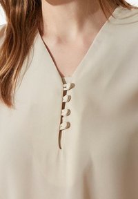 Beige Bluse mit V-Ausschnitt und fünf dekorativen Knöpfen auf der Vorderseite. Weiche, glatte Textur mit lässiger Passform.