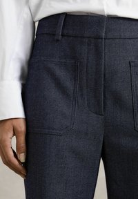 Pantaloni din denim albastru închis, cu o textură netedă, având două buzunare la spate și o croială dreaptă. Asortați cu o cămașă albă cu mânecă lungă.