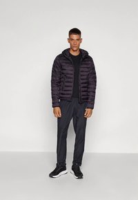 Melnā pufa jaka ar horizontālām šuvēm un kapuci, kopā ar melnām biksēm un melnām sporta kurpēm. Apģērbs ir ikdienišķa stila.