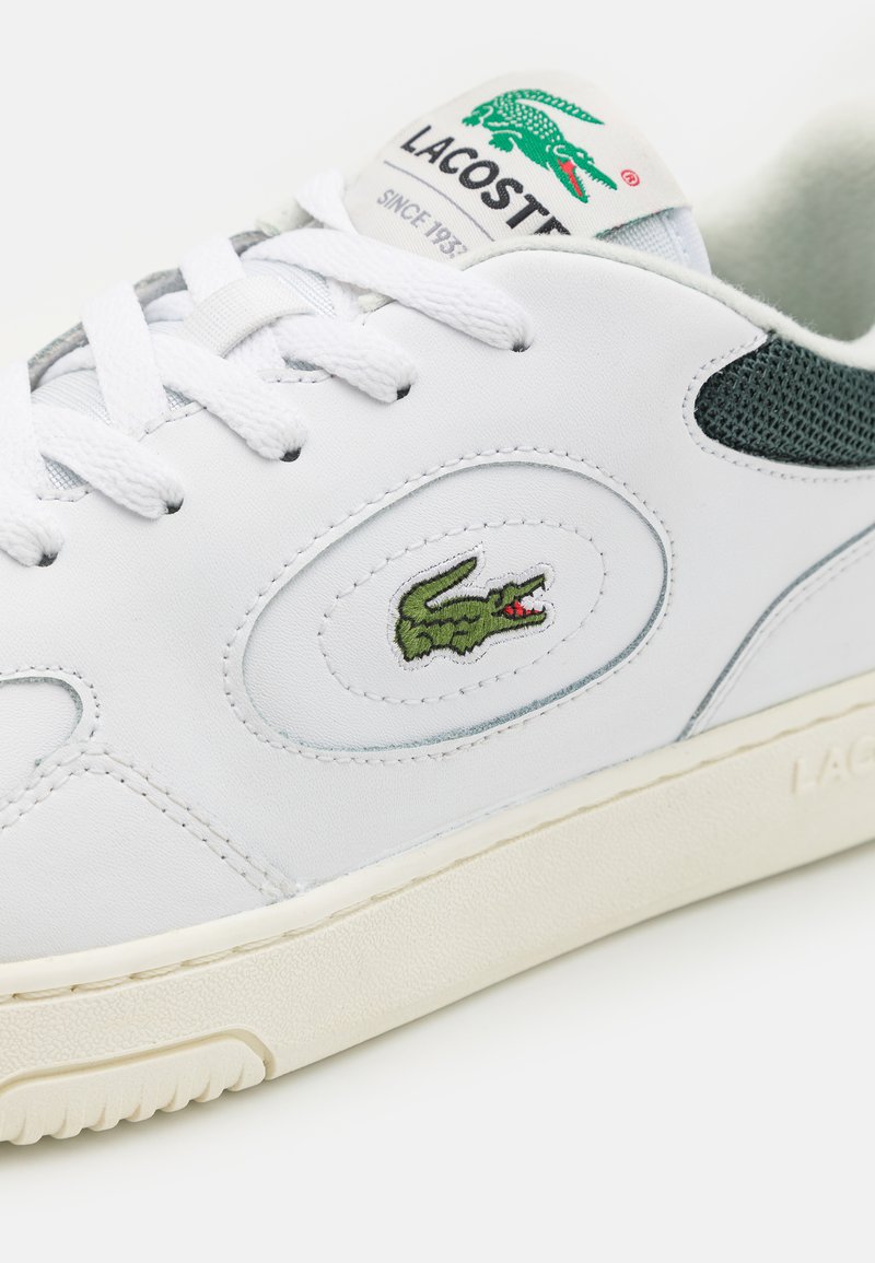 Sneaker bianca con pelle liscia, dettagli in rete verde e logo di coccodrillo verde. Suola testurizzata con una leggera arcata.