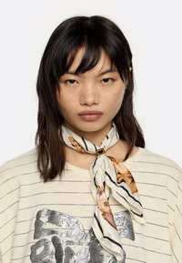 Jedwabny szal w wzór panterki w ciepłych odcieniach, zawiązany na szyi. Beżowa koszula w paski z metalicznym nadrukiem pod spodem.