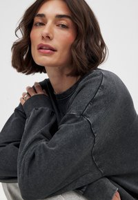 Zwarte oversized sweatshirt van textuurstof, met verlaagde schouders en geribbelde manchetten. Persoon poseert met over elkaar geslagen armen, waardoor de pasvorm van het kledingstuk goed zichtbaar is.