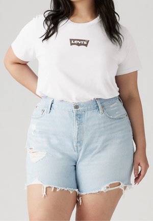 Person iført en hvid Levi’s T-shirt og lyseblå, højttaljede denimshorts med slid og flossede kanter står mod en ensfarvet baggrund.