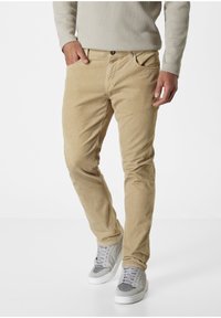 Beige Cordhosen mit schmaler Passform, ausgestattet mit Fronttaschen und einem Knopfverschluss. Kombiniert mit hellgrauen Sneakers für einen lässigen Look.
